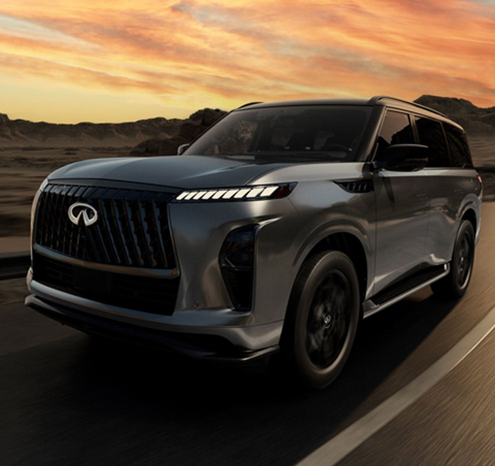 A dark gray Infiniti SUV with sleek headlights drives on a highway toward rocky mountains under an orange sunset.